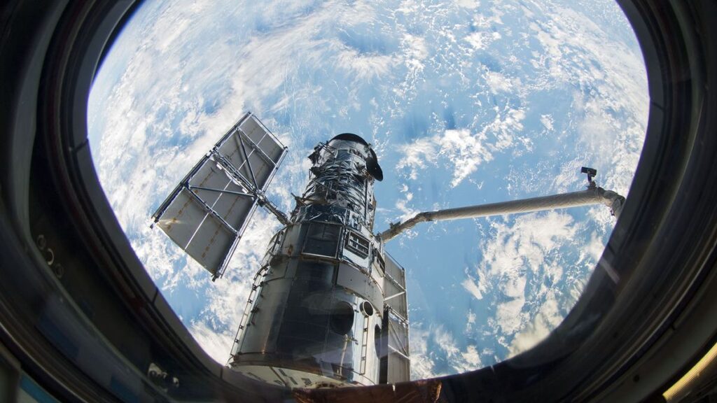 a view of a telescope and robotic arm through the window of a space shuttle. earth and clouds are in the background