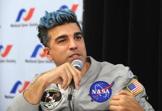 a man with a mohawk in a nasa t-shirt speaks into a microphone while sitting at a table on a stage