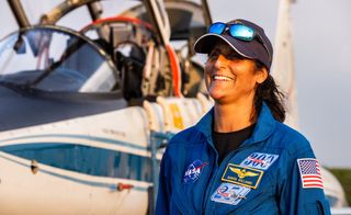 smiling female astronaut in a flight suit and hat in front of a jet plane, with open cockpit