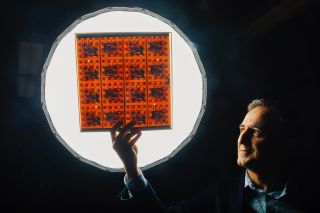 a man against a dimly lit wall holds a transparent sheet with an orange-colored grid in front of a well-lit circle.