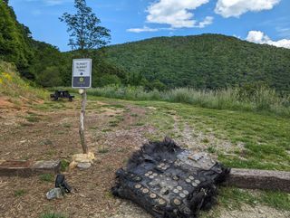 a large black piece of fiberglass covered in bolts and metal plates lies on the ground next to a path leading into a forest.  we can see the mountains rolling in the distance