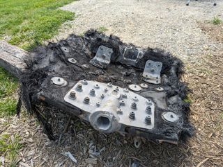 a large black piece of fiberglass covered in bolts and metal plates lies on the ground next to a path leading into a forest.  we can see the mountains rolling in the distance