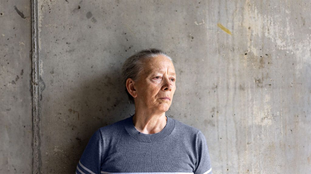 A pensive older woman stands against a gray wall