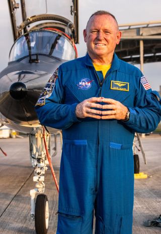 an astronaut in a flight suit touching his hands in front of his chest.  behind him is a jet with open cockpit