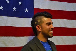 A man wearing a mohawk haircut stands in front of an American flag