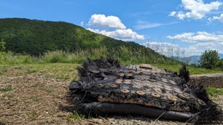 a large black piece of fiberglass covered in bolts and metal plates lies on the ground next to a path leading into a forest.  we can see the mountains rolling in the distance