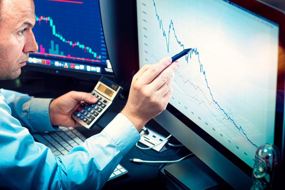 A money manager using a calculator and pen to analyze a stock chart displayed on a computer screen.