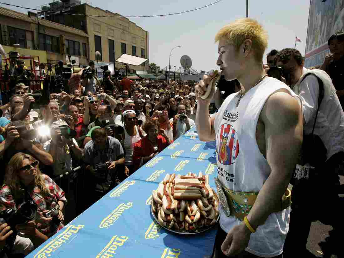 Kobayashi holds a hot dog while photographers in a large crowd take pictures.