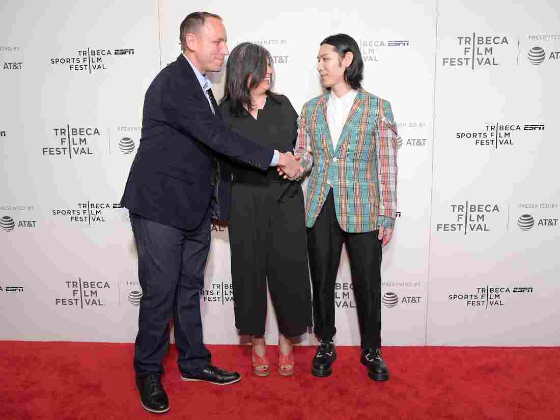 Chestnut and Kobayashi shake hands on a red carpet, with a woman standing between them.