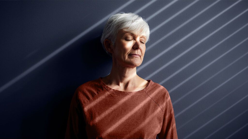 photo of older white woman with white cropped hair standing with her eyes closed against a dark wall