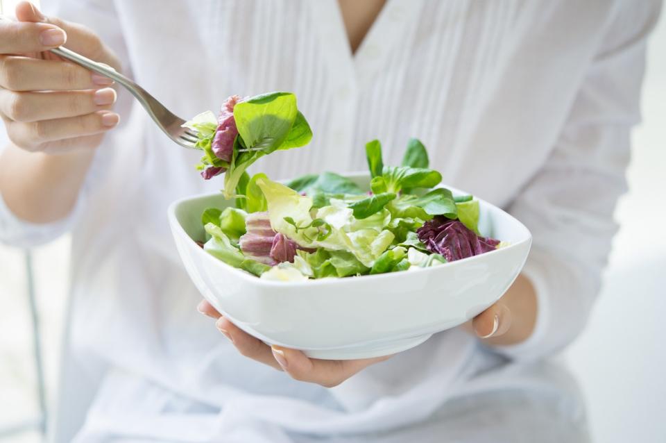 Close-up of a woman holding a plate of fresh green salad in the beautiful morning light.  She is holding a fork and about to eat vegetarian food.  Healthy food and diet concept.  Shallow depth of field with focus on the fork.