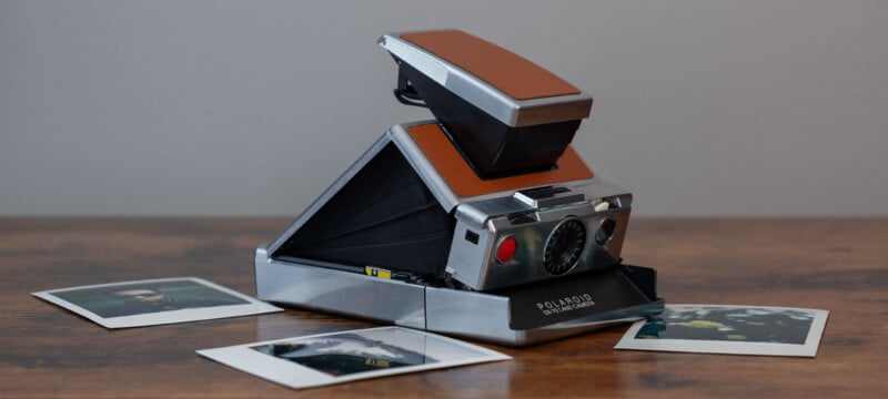 A vintage Polaroid SX-70 Land camera with a brown leather exterior sits on a wooden surface.  The camera is partially unfolded.  Two snapshot photographs are scattered in the foreground, partially visible.  The background is a plain gray wall.