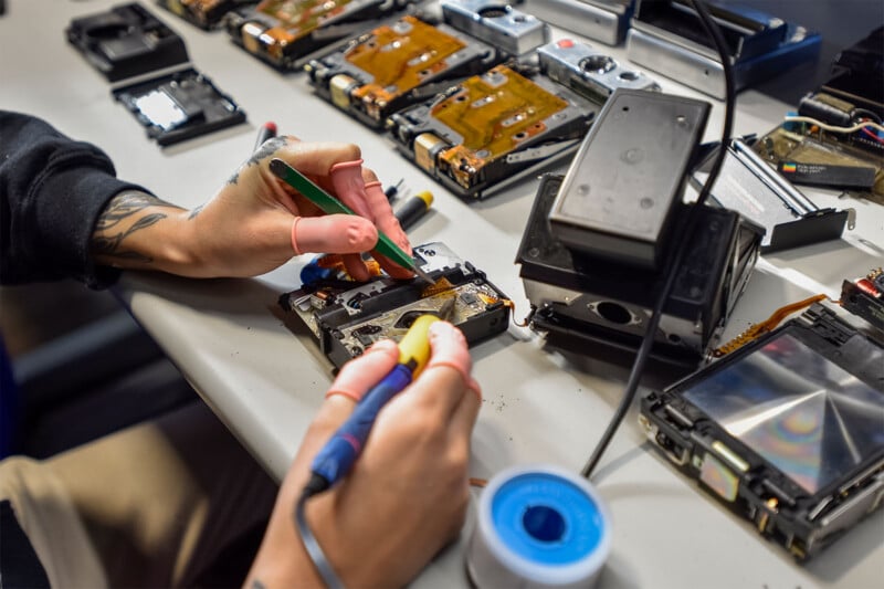 A person's hands are seen working on a table filled with electronic components and tools.  One hand uses a soldering iron while the other holds a green tool.  Various disassembled devices and parts, including hard drives, are distributed throughout the workspace.