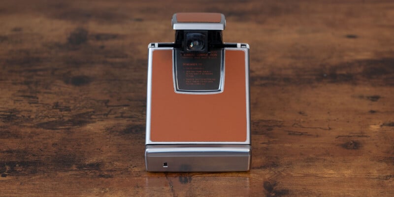 A vintage Polaroid SX-70 Land camera with a brown leather exterior and silver metal frame sits on a wooden surface.  The camera is in the closed position, showing off its rectangular shape and sleek design.