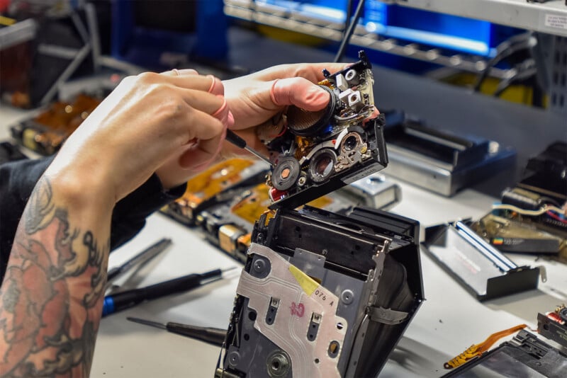 A person with tattooed arms takes apart a camera, with various small tools and electronic components scattered across the workbench.  The background includes shelves containing more electronic equipment and a computer monitor.