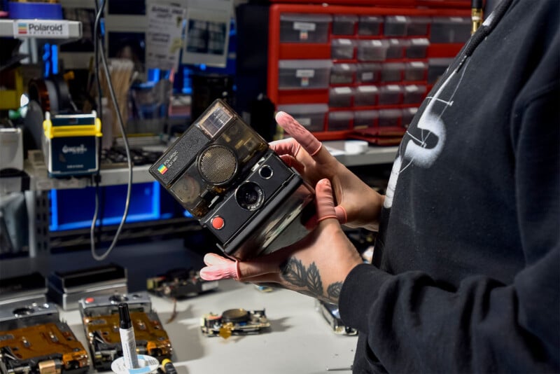 A person holding a vintage Polaroid camera in a workshop.  The background features various electronic components, tools and storage drawers, indicating a repair or restoration environment.  The person's hand tattoo and a partial view of their sweatshirt are visible.