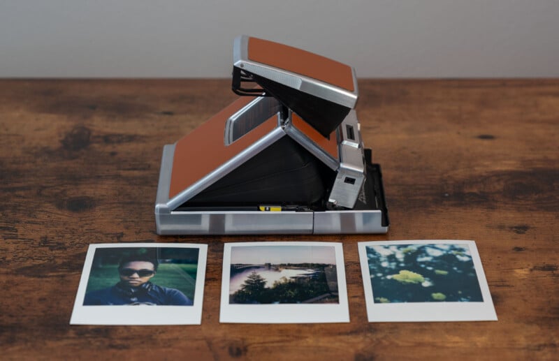 A vintage instant camera with a brown exterior sits on a wooden surface.  In front of him are three Polaroid photos: one of a person wearing sunglasses, one of a panoramic view with a body of water, and one of yellow flowers in the foreground.