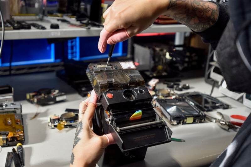 A person with tattooed arms uses a screwdriver to repair or assemble a vintage Polaroid camera at a workstation.  Various electronic components and tools are scattered on the white desk around the workspace.