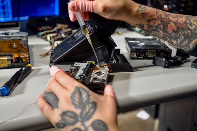 A person with tattooed hands and pink gloves repairs electronic components at a workstation.  They use a syringe to apply liquid to a part while various tools and electronic devices are spread out on the table.