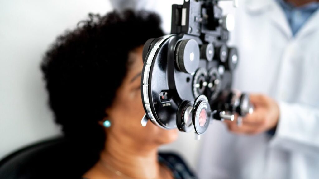 Mature woman is shown having her eyes tested by an opthalomogist. A black device is in front of her eyes which is in focus. The rest of the image is blurred