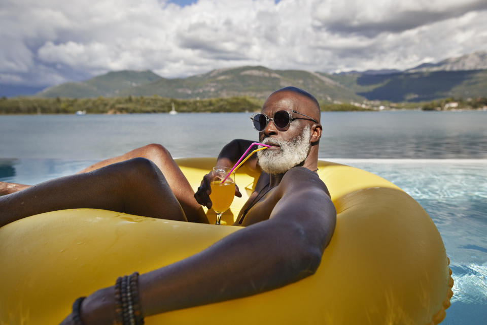 A man with a white beard and sunglasses relaxes on a yellow float in a swimming pool while drinking an orange drink through a colorful straw, with mountains in the background