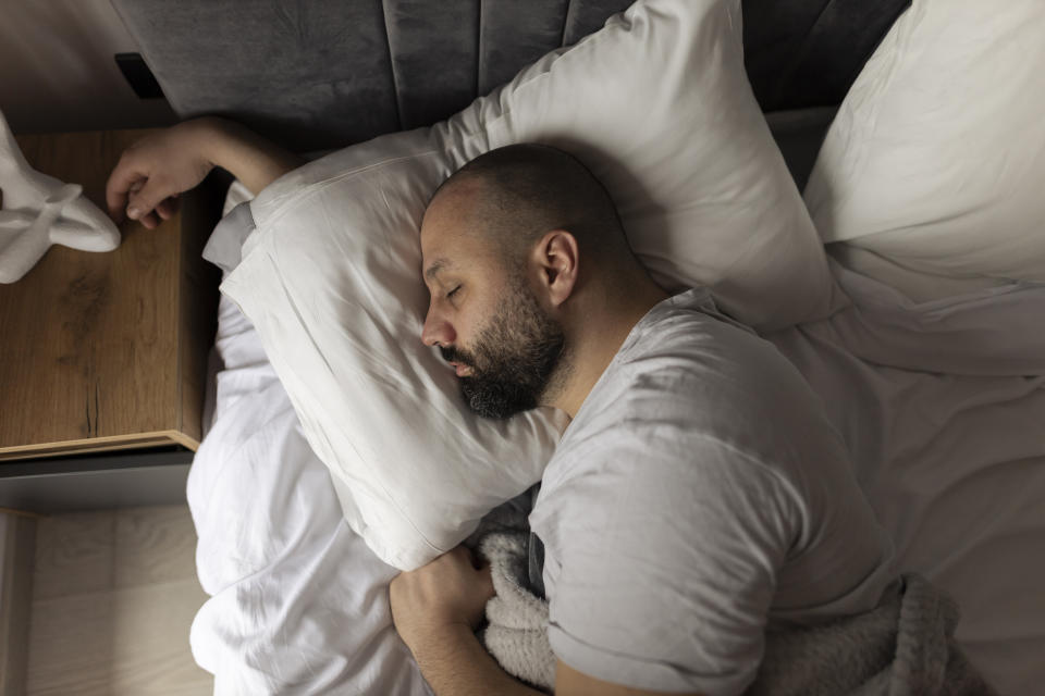 A bearded man with a shaved head sleeps peacefully on a white pillow in a cozy bed.  He is wearing a white t-shirt and has a calm expression