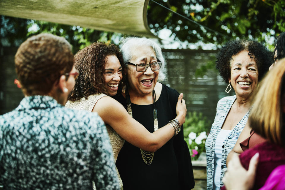 A group of women, including an embracing elderly woman, are outside under an awning, smiling and engaging in cheerful conversation.