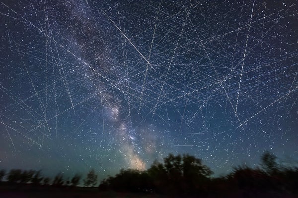 Long exposure composite photograph showing streaks of light from satellites in low Earth orbit filling the sky, a treeline is silhouetted at the bottom of the frame