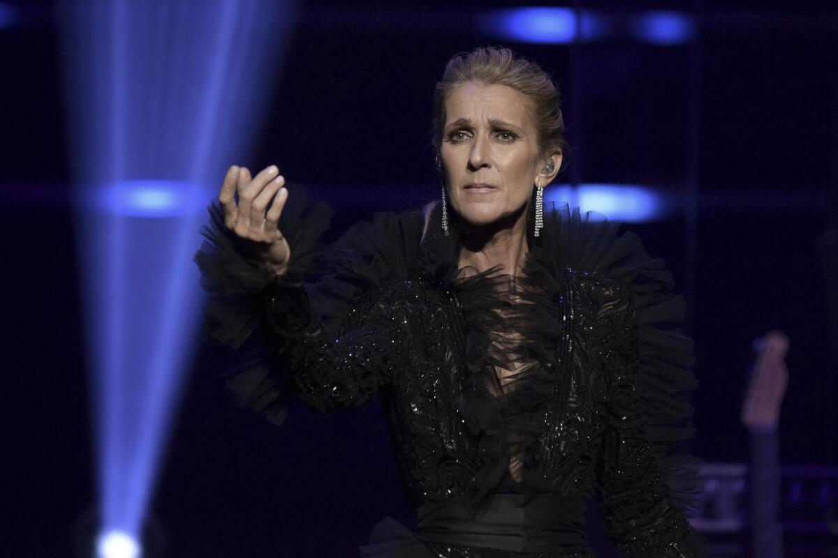 Celine Dion standing on a stage backlit by blue lights in a black outfit holding her arm in front of her.