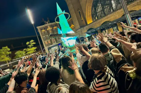 Square Enix Dozens of people form a circle around a giant, glowing replica of a teal-colored crystal. Everyone in the crowd has an arm outstretched toward the crystal, palm open and fingers spread.