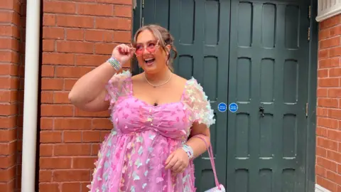 A woman wearing a pink dress and sunglasses smiling and looking to the left 