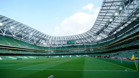 PA An empty Aviva Stadium in Dublin, visible when no concerts or sporting events are taking place. Three-quarters of the stadium and three rows of green seats surrounding a lush football pitch can be seen. 