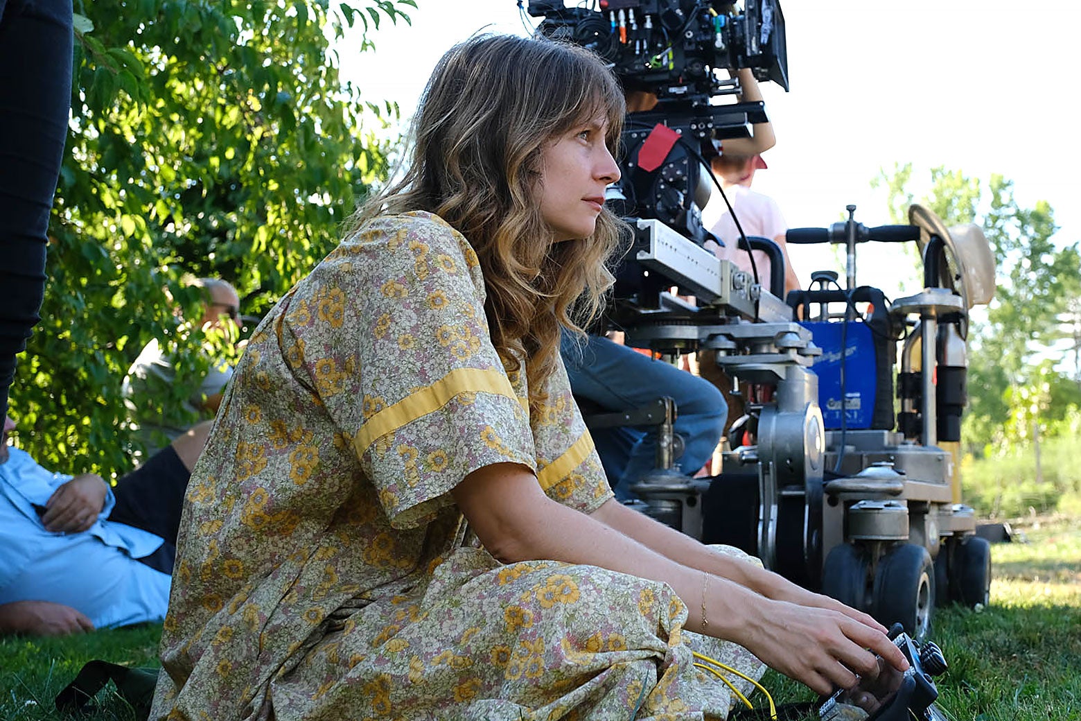 A woman in a floral print dress sits on a grass while a camera films next to her.