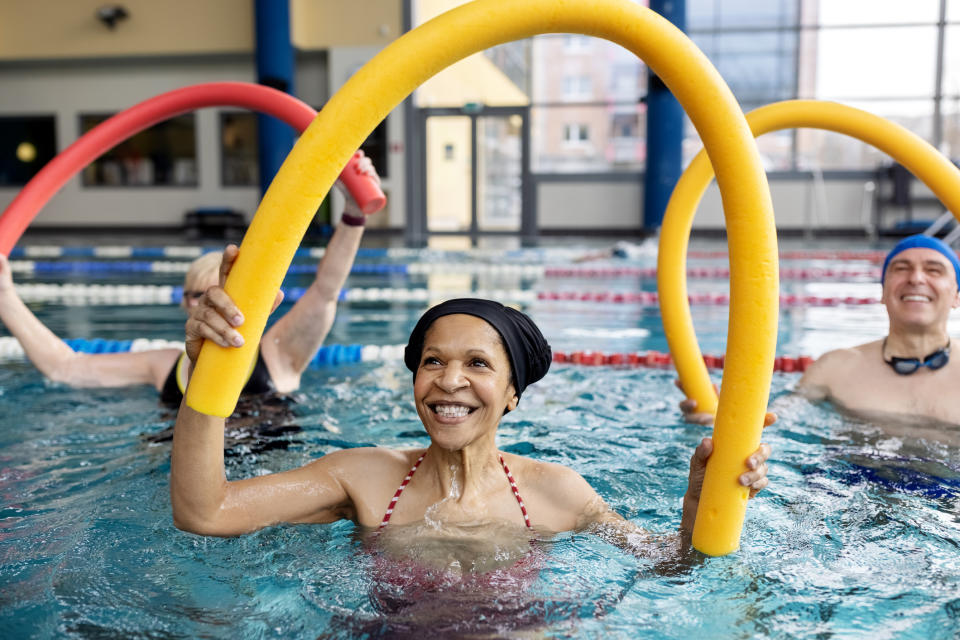 Several smiling people in a swimming pool use pool noodles for exercise.  The focus is on a woman in a swimsuit and swimming cap, holding a yellow noodle above her head.