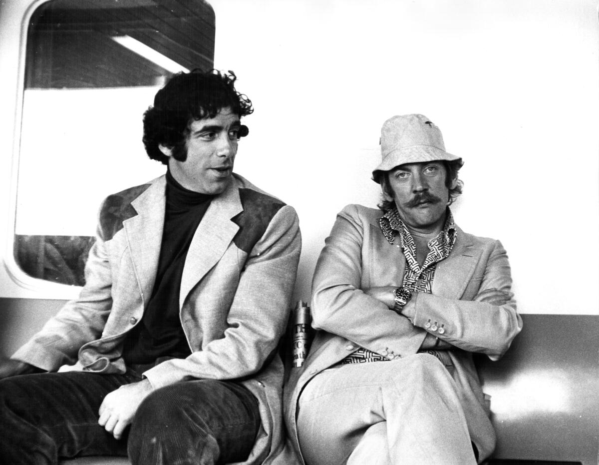 Elliott Gould looks at Donald Sutherland with his arms crossed as they both sit on a sofa.