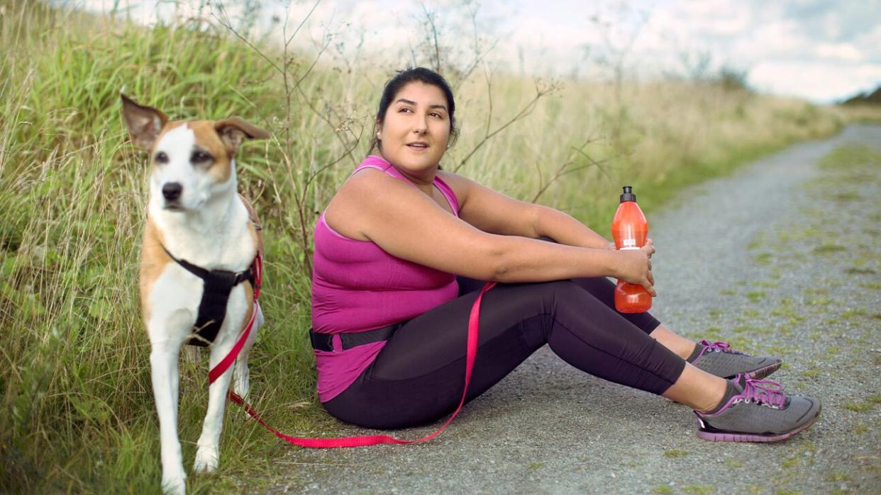 A woman in sportswear takes a break on a road next to a white and brown dog.