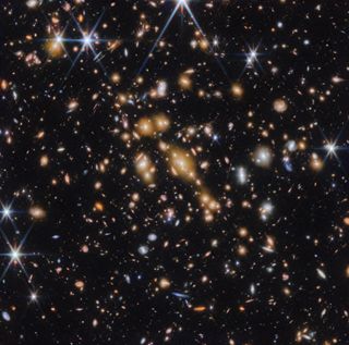 A field of galaxies against the black background of space.  In the middle is a collection of dozens of yellowish galaxies that form a galaxy cluster in the foreground.  Among them are distorted linear elements, which mostly appear to follow invisible concentric circles curved around the center of the image.  Linear features are created when light from a background galaxy is bent and magnified by gravitational lensing.  A variety of brightly colored red and blue galaxies of various shapes are scattered across the image, giving it the appearance of being densely populated.