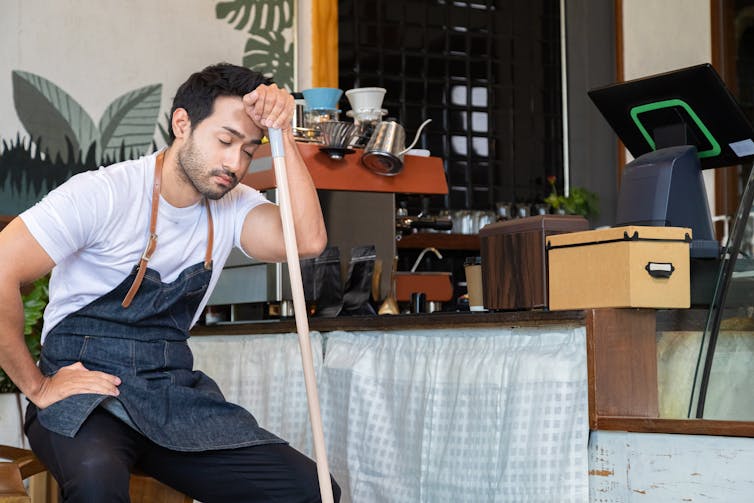 Man wearing an apron, sitting, resting his forehead on the end of a broom, looking tired.