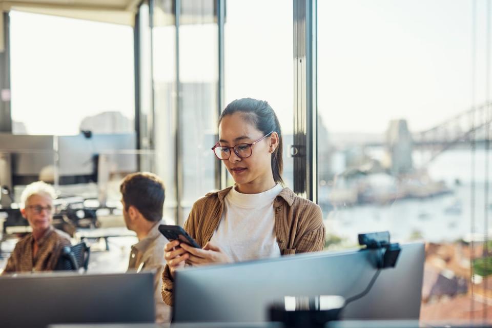 A person standing in an office looks at their cell phone in front of two computer screens. 