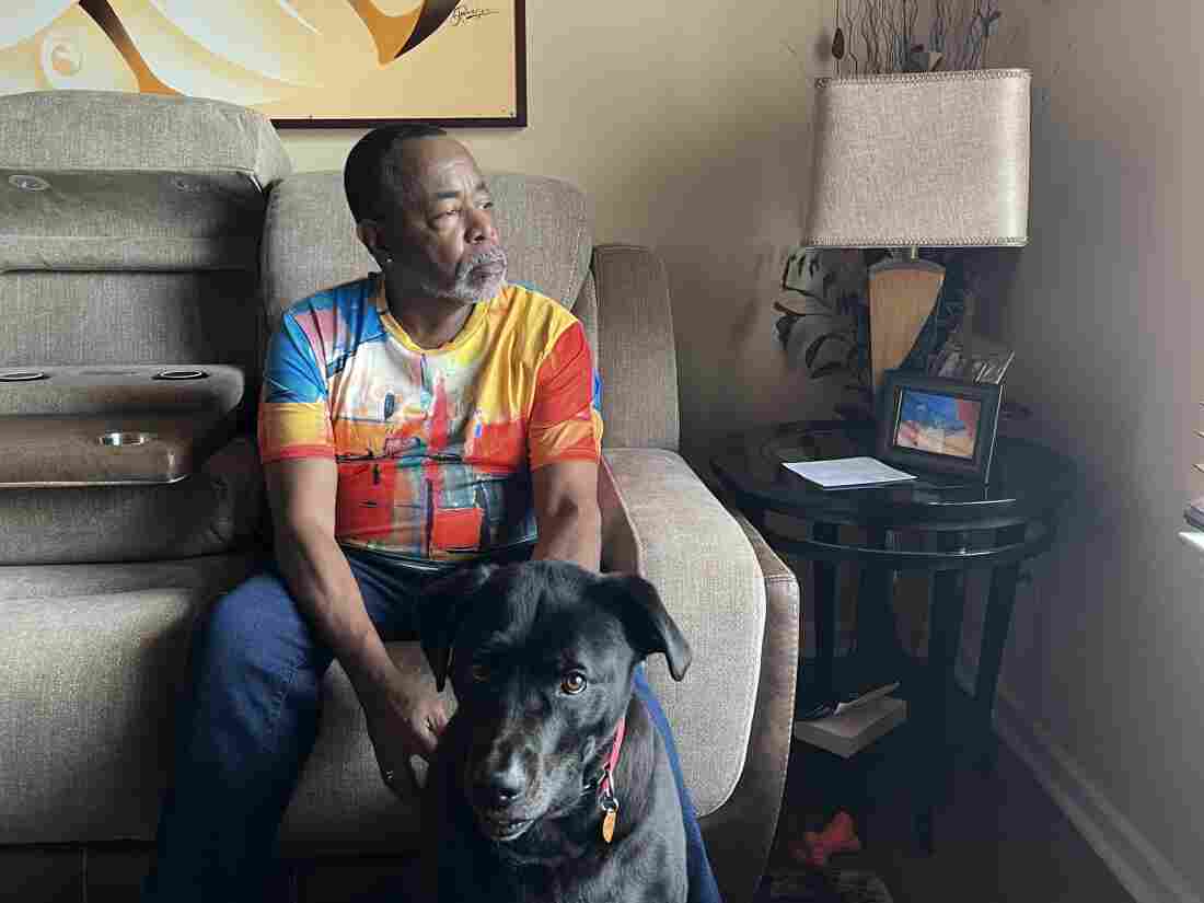 A man in a colorful shirt with a gray beard, Malcolm Reid, sits on his beige sofa in the living room.  Reid's hands are on his dog, Sampson, a black Labrador mix sitting in front of the couch.  The dog looks at the camera while Reid looks towards a window. 