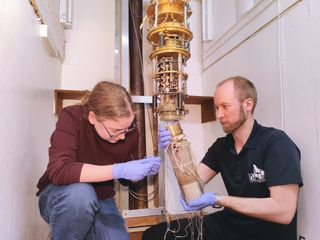 A white room containing two crouched people tending to a complex gold machine