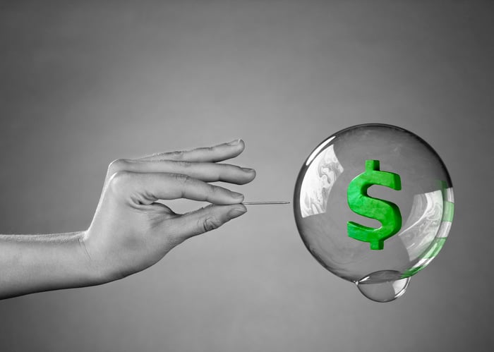 A person uses a pin to pop a bubble containing a green dollar sign.