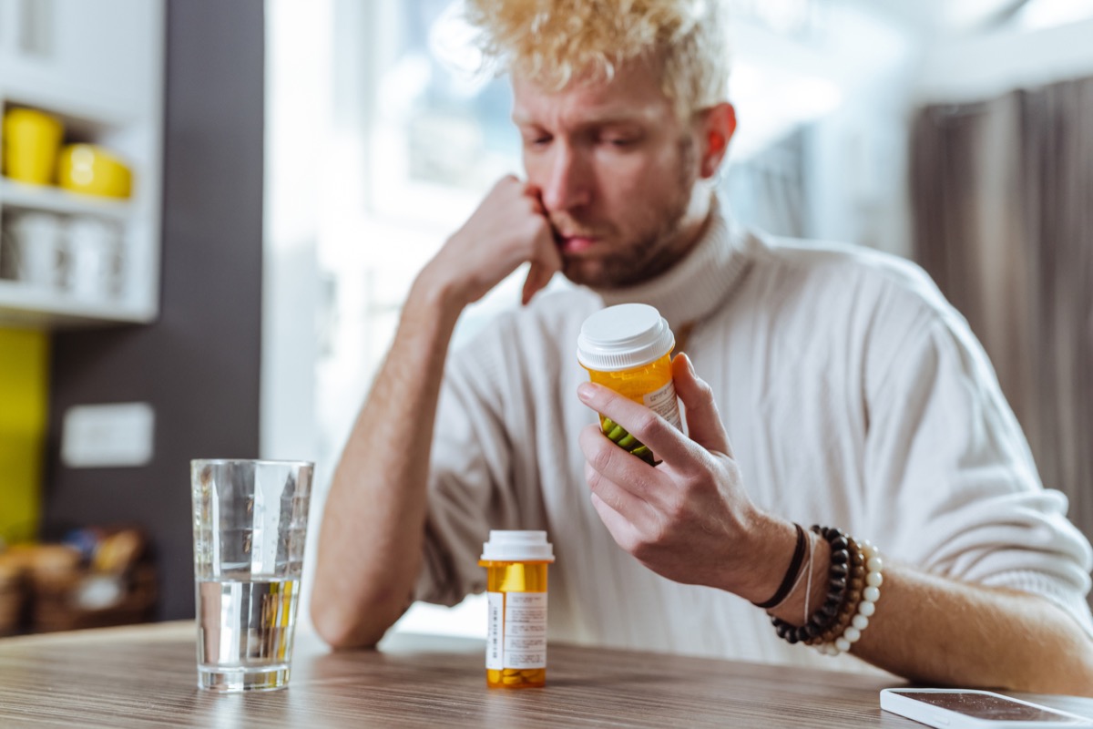 A man sits and holds his head in his hands while thinking and looking at pill bottles