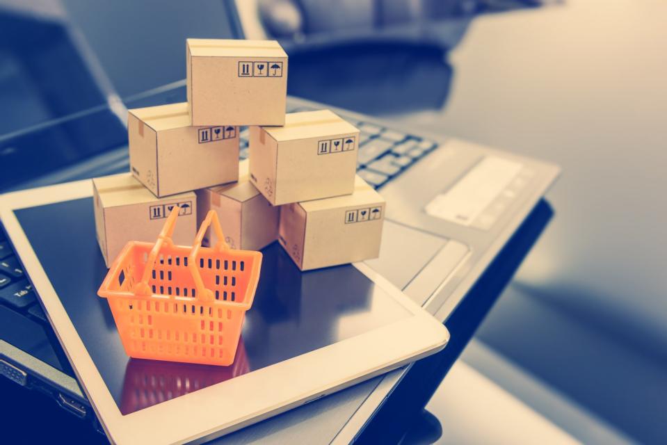 A miniature orange hand basket and a mini pyramid of boxes resting on a tablet and an open laptop. 