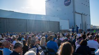 Two men in the distance stand in front of a large building that blocks the sun. It bears the NASA logo near the top. They address a large crowd, stretching out the rest of the frame.