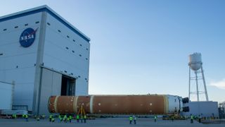 A large orange rocket booster stage sits horizontally on wheels outside a large hangar and water tower.