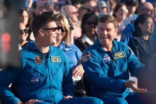 Two men in blue jumpsuits sit laughing in a crowd.