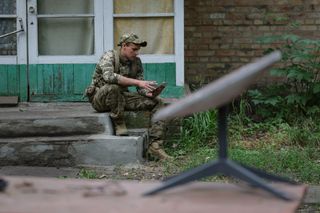 A soldier in camouflage sits on a porch and looks at a cell phone behind a small portable satellite dish