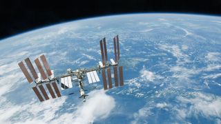 A T-shaped space station with multiple solar panels floats above Earth