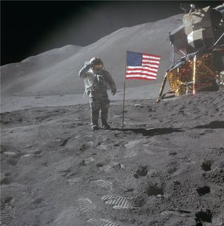 An astronaut in a bulky white spacesuit plants an American flag on the gray, dusty surface of the moon